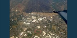 Aerial view of a university campus with a red running track and numerous white buildings, mountains in the background, and part of an airplane wing on the right side.
