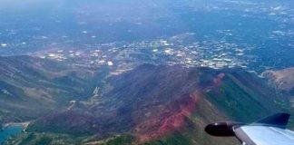 Aerial view from a small plane, wingtip visible on the right, over green and brown mountains with a distant town in the valley.