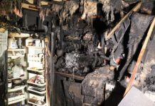 Extensively burned room with charred debris, an open refrigerator door showing shelves, and soot-covered walls from a fire.