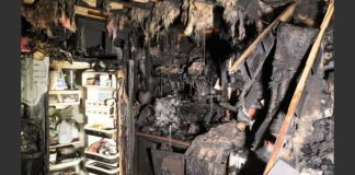 Extensively burned room with charred debris, an open refrigerator door showing shelves, and soot-covered walls from a fire.