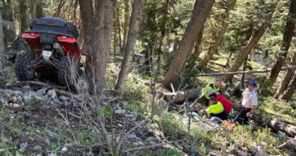 Red ATV stuck on a rocky slope in a forest, with three people in bright vests helping to pull it free.