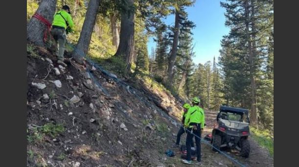 Two workers in bright safety jackets stand on a rocky forest slope near a utility vehicle, with ropes and gear laid out for stability or rescue work.