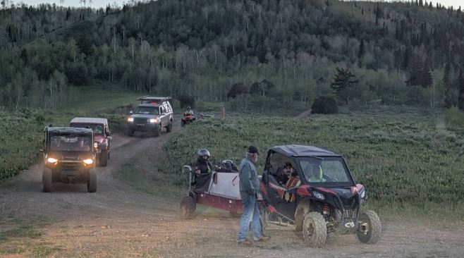 Group of off-road vehicles driving along a dirt path through a grassy valley with forested hills in the background; a man in blue jeans and helmet stands near a red side-by-side.