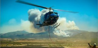 Blue helicopter hovering low over a desert airstrip, rope extending to a red bag on the ground as a worker in a helmet signals nearby, with smoke rising in the background mountains.