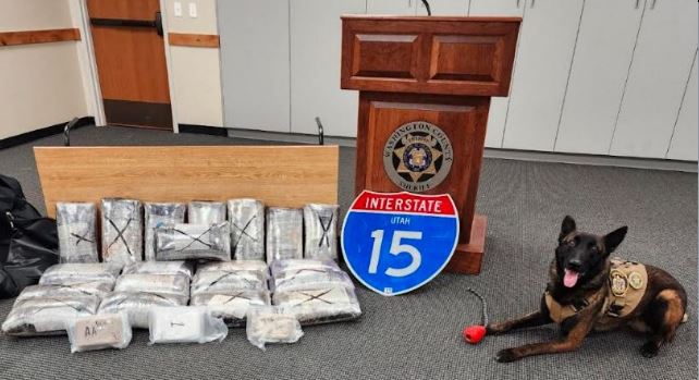 Press podium with seal, blue Utah I-15 sign, and bundles wrapped in plastic on the floor next to a police dog in a vest.