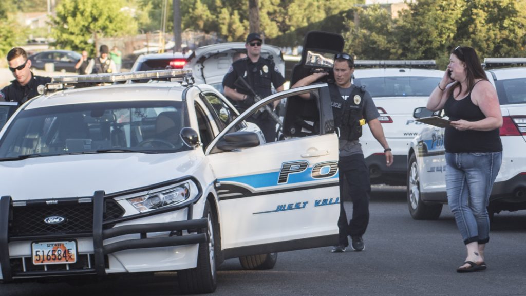 Police officers stand near a white patrol car with blue graphics, a civilian woman talking on her phone nearby in a daytime street scene.