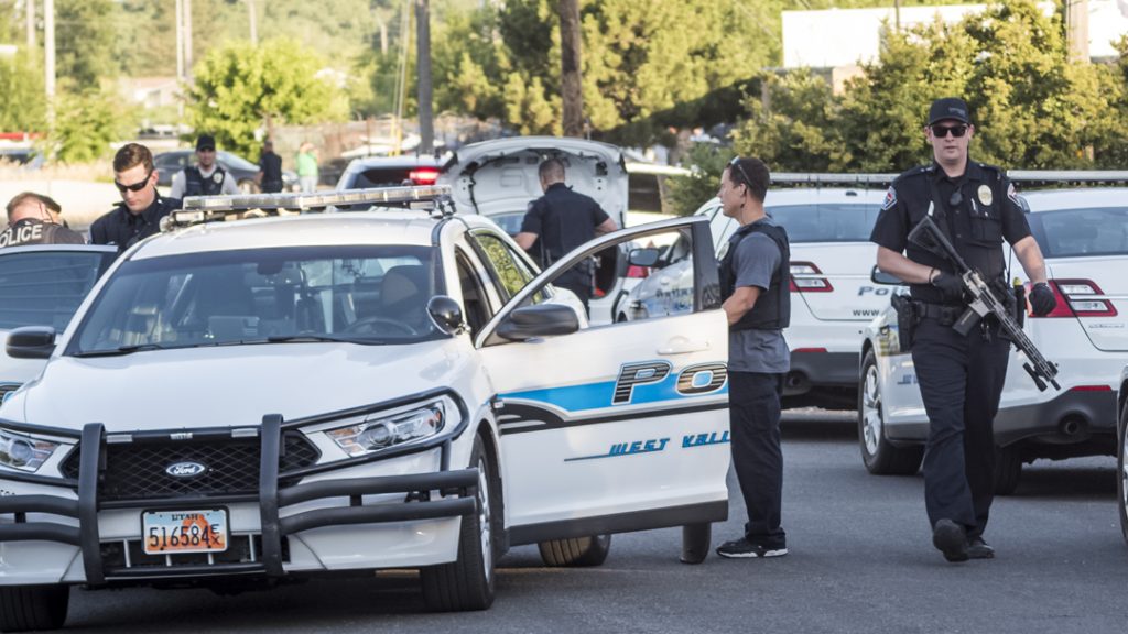 White police SUV with blue stripe, several officers in tactical gear stationing a scene, one officer carrying a rifle as others stand nearby near more patrol cars in the background.