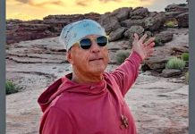 Man wearing a light blue bandana and sunglasses, red hoodie, arms outstretched in a rocky desert at sunset.
