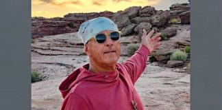 Man wearing a light blue bandana and sunglasses, red hoodie, arms outstretched in a rocky desert at sunset.