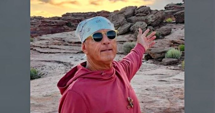 Danny Joe Kregle Man wearing a light blue bandana and sunglasses, red hoodie, arms outstretched in a rocky desert at sunset.