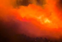 Large fiery blaze at night with bright orange flames and dark smoke rising above a silhouetted landscape.