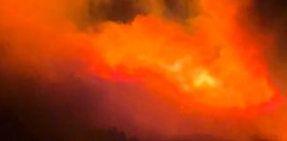 Large fiery blaze at night with bright orange flames and dark smoke rising above a silhouetted landscape.