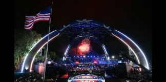 Nighttime outdoor concert under a large illuminated arch stage with fireworks, a cheering crowd, and an American flag waving on the left.