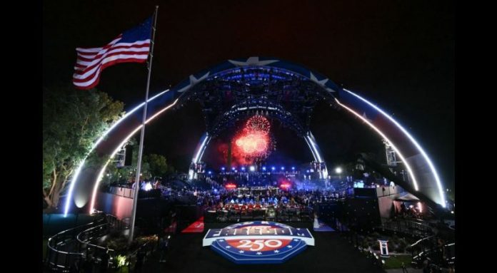 Nighttime outdoor concert under a large illuminated arch stage with fireworks, a cheering crowd, and an American flag waving on the left.