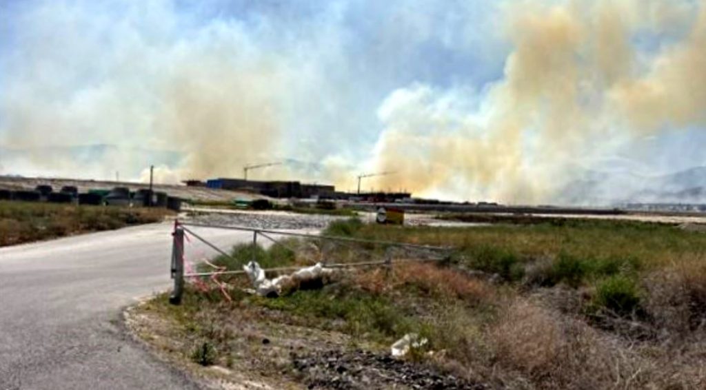 Roadside scene with a large plume of smoke rising from distant buildings; open gate and dry scrub in the foreground.
