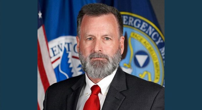 Gregg_Phillips_FEMA Man in a dark suit and red tie with a gray beard, posed in front of emergency management seals and flags.