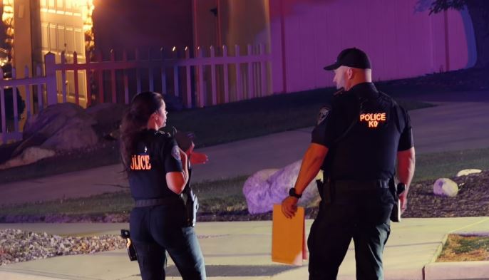 Two police officers stand outside at night, talking near a sidewalk and a lit house behind a fence.
