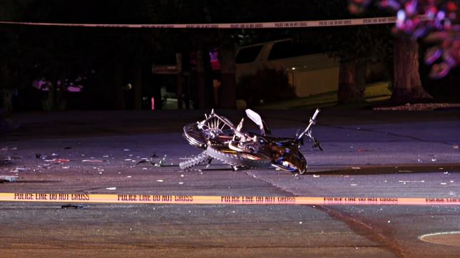 Motorcycle crash scene at night with a damaged bike lying in the street, debris scattered around, and yellow police tape in the foreground.