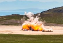 Explosion in a barren landscape with orange flames and white smoke rising from ground blast site, hills in background.