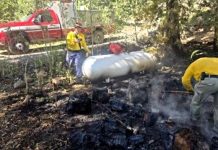 Firefighters extinguish a forest fire around a large propane tank; smoke rises as a red fire truck sits in the background.