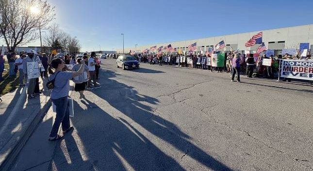 ICE-facility-protest Crowd of protesters and supporters line a sunny street with American flags and banners; a car drives down the road while onlookers watch from the sidewalk.