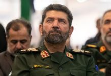 Middle-aged man in a green military uniform with a beard, seated among others at an indoor event.