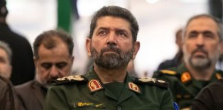 Middle-aged man in a green military uniform with a beard, seated among others at an indoor event.
