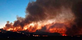 Wildfire on a landscape at dusk with bright flames and thick black smoke rising into the sky, casting an orange glow over the scene