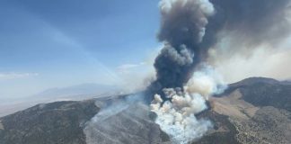 Tall smoke plume rising from a wildfire on a hillside in a mountainous area, with scorched terrain nearby.