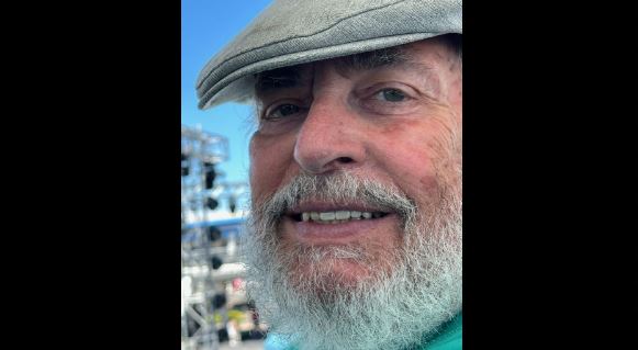Close-up of an older man with a gray beard and a flat cap, smiling outdoors with a bright blue sky in the background.