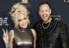 Smiling blonde woman in a shiny sleeveless dress makes a peace sign beside a tattooed man in a black patterned jacket at an awards event.