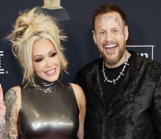 Smiling blonde woman in a shiny sleeveless dress makes a peace sign beside a tattooed man in a black patterned jacket at an awards event.