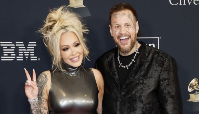 Smiling blonde woman in a shiny sleeveless dress makes a peace sign beside a tattooed man in a black patterned jacket at an awards event.