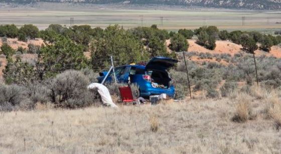 Blue hatchback with trunk open, parked in a dry, brushy field as a small camping setup unfolds nearby.