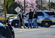 Police officers stand in a group near a marked sheriff/patrol SUV behind yellow crime-scene tape on a street.