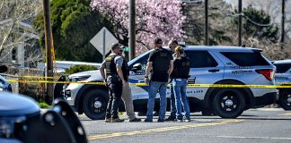 Police officers stand in a group near a marked sheriff/patrol SUV behind yellow crime-scene tape on a street.