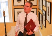 A middle-aged man in a white shirt and brown tie walks down a hallway lined with framed photos, holding a red folder.