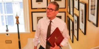 A middle-aged man in a white shirt and brown tie walks down a hallway lined with framed photos, holding a red folder.
