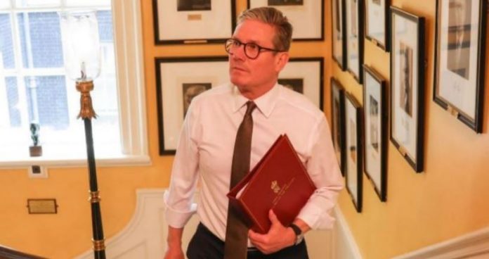 Keir Starmer A middle-aged man in a white shirt and brown tie walks down a hallway lined with framed photos, holding a red folder.
