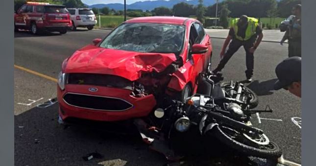 Red car with a crumpled front after colliding with a black motorcycle; responders and bystanders at the scene on a roadway.