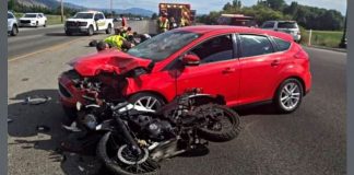 Red hatchback with severe front-end damage after a collision with a black motorcycle; emergency responders at the scene on a highway.