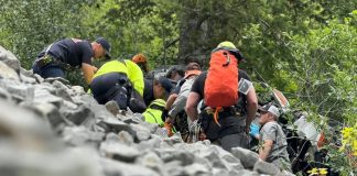 Group of hikers and rescuers climbing a steep rocky slope in a forest, with backpacks and safety gear visible.