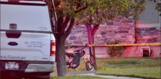A bicycle leaning against a tree beside yellow police tape on a residential street, with an emergency vehicle parked nearby.