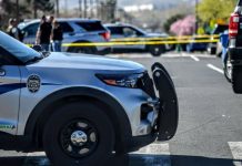 Front of a police SUV with a bull bar, parked at a taped-off street scene; yellow police tape and other vehicles in the background.