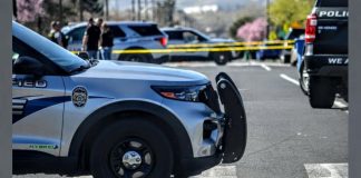 Front of a police SUV with a bull bar, parked at a taped-off street scene; yellow police tape and other vehicles in the background.