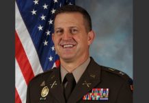 Smiling male U.S. Army officer in dress uniform with ribbons, standing before the American flag.