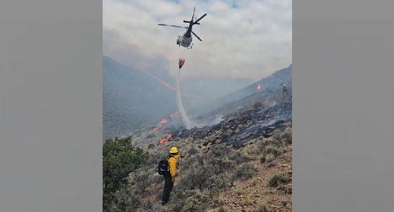 Helicopter dropping water onto a hillside wildfire as a firefighter in yellow gear stands on rocky terrain watching.