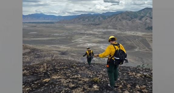 Two hikers in yellow jackets descend a rocky slope toward a valley and distant mountains, wearing backpacks.