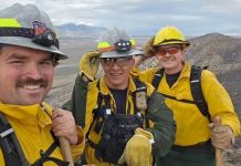 Three outdoor workers in yellow jackets and helmets pose for a selfie on a rocky hillside with a canyon backdrop.
