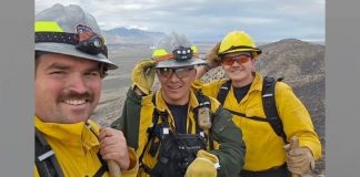Three outdoor workers in yellow jackets and helmets pose for a selfie on a rocky hillside with a canyon backdrop.
